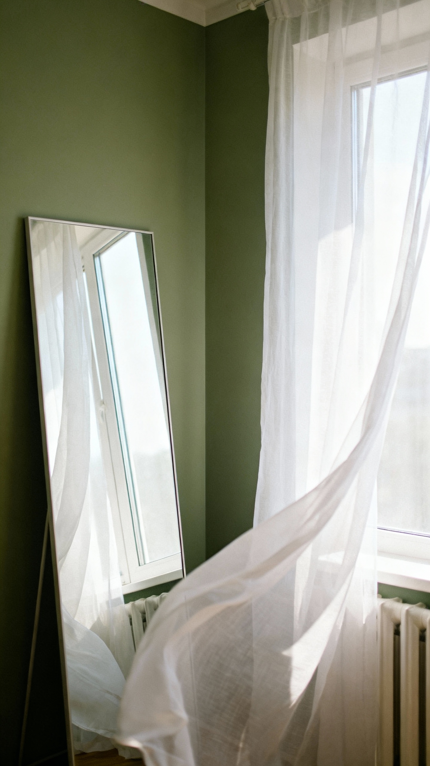 Large floor mirror opposite window reflecting light, sage walls, sheer curtains